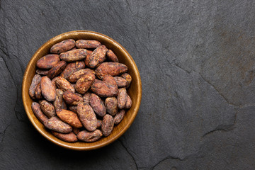 Roasted unpeeled cocoa beans in a brown wooden bowl isolated on black slate from above. Space for...