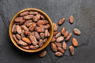 Roasted unpeeled cocoa beans in a brown wooden bowl next to unpeeled cocoa beansl isolated on black slate from above.