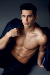 Muscular, young brunette man sitting and posing in fashionable suit on a naked torso, isolated on...