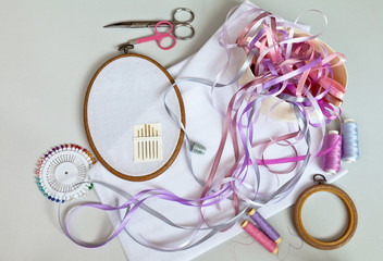 Needlework. Accessories for embroidery with satin ribbons: needles, embroidery frames, colored ribbons, canvases, scissors, thread, pins and thimble on light table. Flat lay, top view