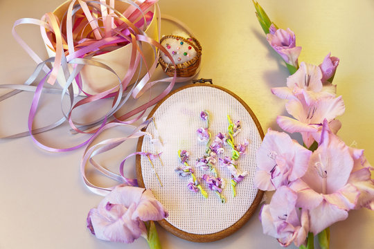 The embroidery process with satin ribbons of light-violet gladiolus flowers on white convas in oval fraime. Top view, sunset light  (Embroidery with satin ribbons were performed by author of images)