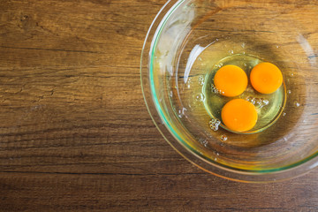 Three raw chicken egg yolk and white in glass bowl on top of wooden table top.	