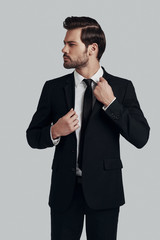 Confidence and charisma. Handsome young man in full suit adjusting jacket and looking away while standing against grey background