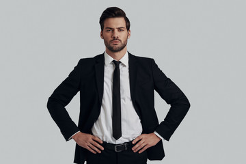 Fototapeta premium Portrait of confidence. Handsome young man in full suit looking at camera and keeping hands on hips while standing against grey background