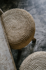 Fototapeta na wymiar Natural straw round pouf cushion chair with wooden legs, grey concrete floor. Close up details. Minimalist loft coffee shop, restaurant, home interior design