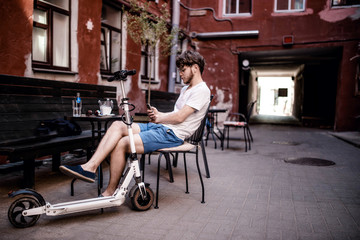 Obraz premium young man with an electric scooter is drinking coffee on the terrace of a cozy cafe in the city. Rent of electric transport.
