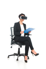 mixed race businesswoman using virtual reality headset while sitting in office chair and holding...