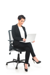 smiling mixed race businesswoman sitting in office chair and using laptop on white background