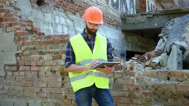 An engineer builder in a helmet is calculating the building demolition plan and looking at the construction project on a tablet, the ruins in the background, middle shot