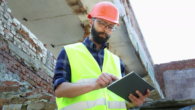 An engineer builder in a helmet use tablet, building demolition plan and looking at the construction project. Destroyed building in the background
