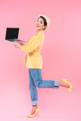 cheerful mixed race woman in bright clothing and straw hat posing at camera with laptop on pink background