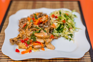 Pieces of rabbit stew with vegetables pepper, onion and herbs with boiled buckwheat and salad of fresh cucumbers and cabbage on a white plate. Mouth-watering delicious food at the gala dinner