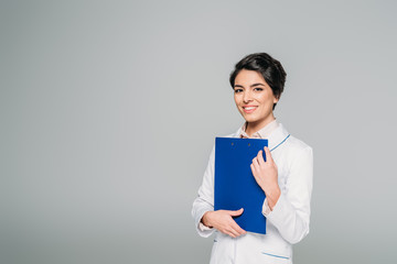 pretty mixed race doctor holding clipboard while smiling at camera isolated on grey