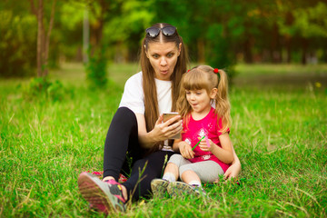 Naklejka na ściany i meble Cheerful smiling happy mother and her little daughter are sitting on green grass in summer park and using a phone for watching cartoons, a girl is indicating on the monitor at the weekend