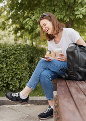 Laughing female sitting on bench. Young cheerful woman relaxing in park with smartphone and paper cup on summer day.