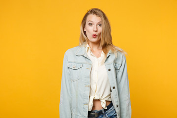 Portrait of amazed surprised young woman in denim casual clothes standing and looking camera isolated on bright yellow orange wall background in studio. People lifestyle concept. Mock up copy space.