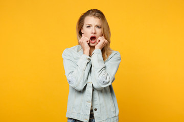 Portrait of shocked scared young woman in denim casual clothes keeping mouth open, putting hands on cheeks isolated on yellow orange background in studio. People lifestyle concept. Mock up copy space.
