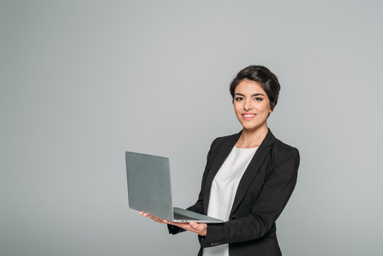 cheerful mixed race businesswoman holding laptop while smiling at camera isolated on grey