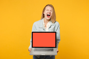 Portrait of wild screaming young woman in denim casual clothes holding laptop pc computer with blank empty screen isolated on yellow orange background. People lifestyle concept. Mock up copy space.