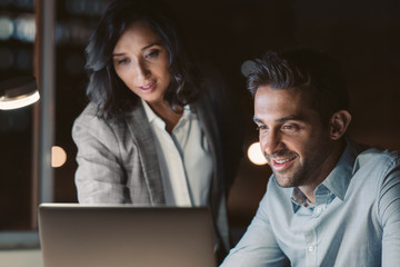 Smiling office colleagues using a laptop while working overtime together