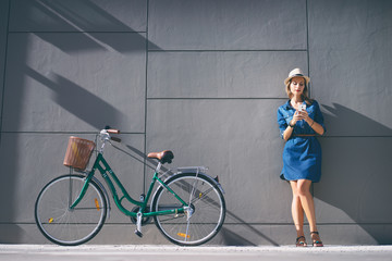 Lifestyle and technology. Young pretty woman using smartphone with bicycle leaning on the grey wall.