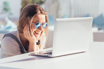 Cheerful beautiful young teenager girl chatting in social networks using a laptop and high-speed Internet while sitting by the sea enjoying the clear sea air and a sunny warm day