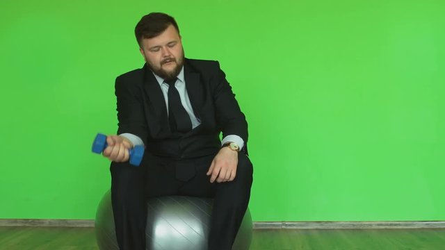 Concept of businessman and fitness. Fat guy with dumbbell sits on Exercise ball and looks at the wrist watch. Plump bearded man in jacket, shirt, tie and trousers prepares to do exercises in the gym