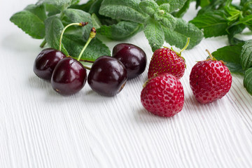 Red cherries, strawberries, green mint and melissa on white wood
