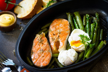 Salmon fish steak grilled with asparagus, poached egg in a frying pan on a rustic stone table. Healthy food.