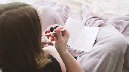 Pregnant woman is sitting on the bed and eating some berries while reading a book. 4k. - Powered by Adobe