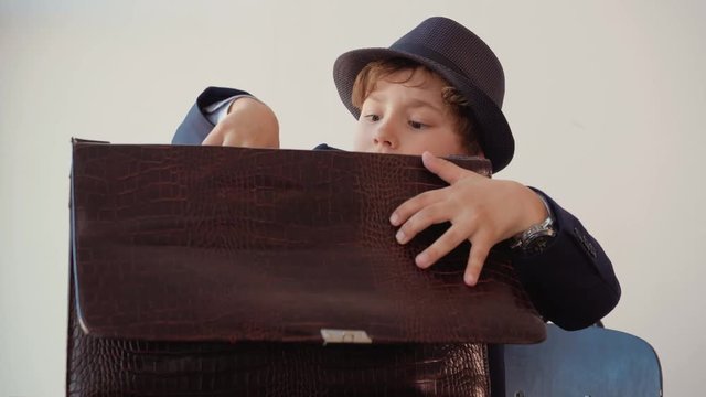 Little boy looks like a businessman in suit and hat takes off a pack of hundred-dollar banknotes out of his briefcase sitting at table in his office. Business and earning money concept.