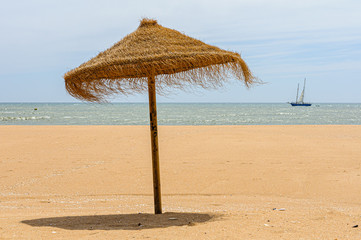 Straw parasol on a sandy beach with a yacht sailing past