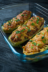 Stuffed zucchini with minced meat and grated cheese in a glass baking sheet. Dark wooden background.