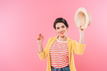 pretty mixed race woman waving straw hat and smiling at camera isolated on pink