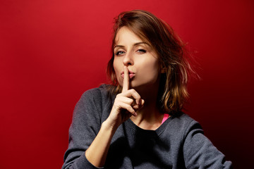 Keep secret! Pretty young woman showing silence sign with her finger on lips. Colorful portrait with red background.