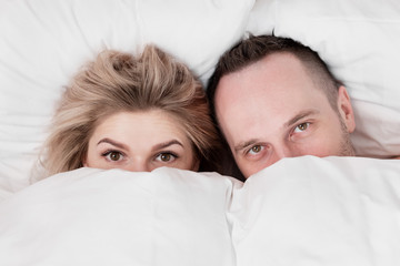 Married couple, man and woman, are lying in bed, hugging and sleeping on white bedding. Hiding their faces under blanket and looking out.