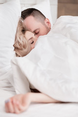 Married couple, man and woman, are lying in bed, hugging and sleeping on white bedding. Hiding their faces under blanket and looking out.