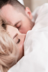Married couple, man and woman, are lying in bed, hugging and sleeping on white bedding. Hiding their faces under blanket and looking out.