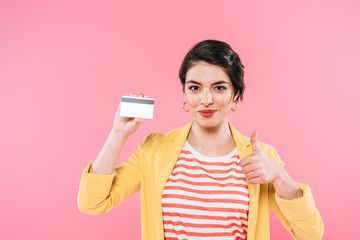 beautiful mixed race woman holding credit card and showing thumb up isolated on pink