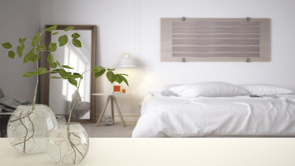White table top or shelf with glass vase with hydroponic plant, ornament, root of plant in water, branch in vase, house plant, white blurred bedroom in the background, interior design