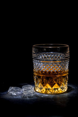 Illuminated crystal glass filled with golden whiskey with lying next to the melted ice cubes on a black background.