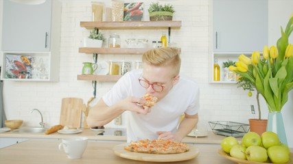 Blonde man is eating pizza while having breakfast at the kitchen