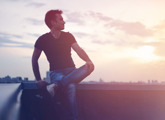 Attractive stylish young man on the roof at sunset looking at t city