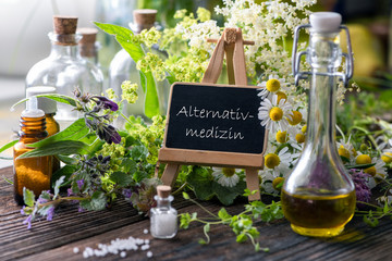 Zusammenstellung verschiedener Heilpflanzen mit Fläschchen und Tigeln und Schild: Alternativmedizin