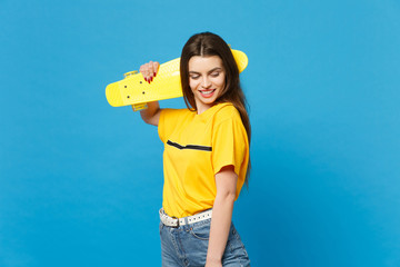 Portrait of pretty smiling young woman in vivid casual clothes standing, looking down, hold yellow skateboard isolated on blue wall background in studio. People lifestyle concept. Mock up copy space.