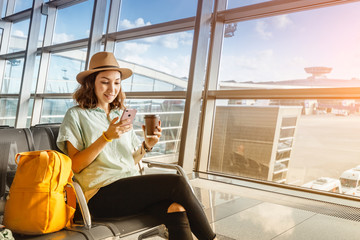 Asian girl waiting for departure at the airport on your vacation. Uses a smartphone and drinks coffee - obrazy, fototapety, plakaty