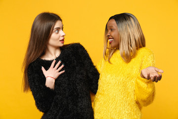 Two young women friends european and african american in black yellow clothes standing posing isolated on bright orange wall background, studio portrait. People lifestyle concept. Mock up copy space.