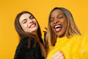 Two women friends european and african american in black yellow clothes doing selfie shot posing isolated on bright orange wall background studio portrait. People lifestyle concept. Mock up copy space