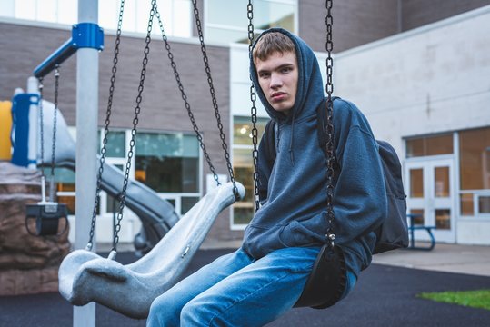 Sad teenager sitting on a swing outside of a school. He is reminiscing about when he was younger.