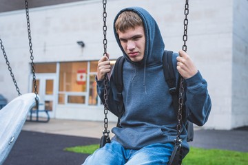 Sad teenager sitting on a swing outside of a school. He is reminiscing about when he was younger.